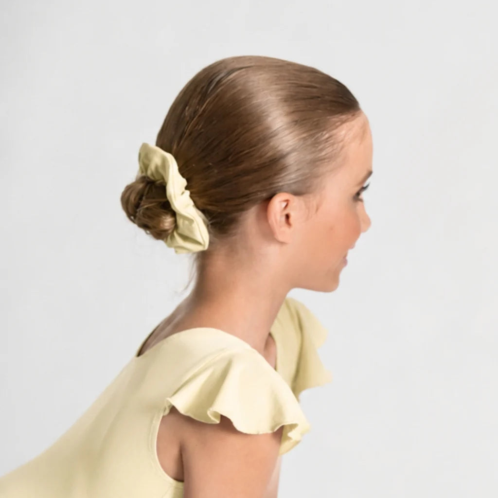 A young girl with light brown hair styled in a low bun, wearing a pale yellow fabric scrunchie. She is shown in profile against a clean, light gray studio background. Ballet Rosa ZOE 854MF